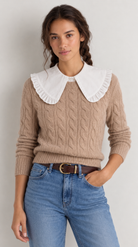 A woman wearing a beige cable-knit sweater with a large white ruffled Peter Pan collar, paired with blue jeans and a brown belt, against a light gray studio background.