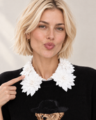 A blonde woman wearing a black knit sweater with an ornate floral collar by the Curated Collar, pointing to the collar with her finger against a light beige background.  