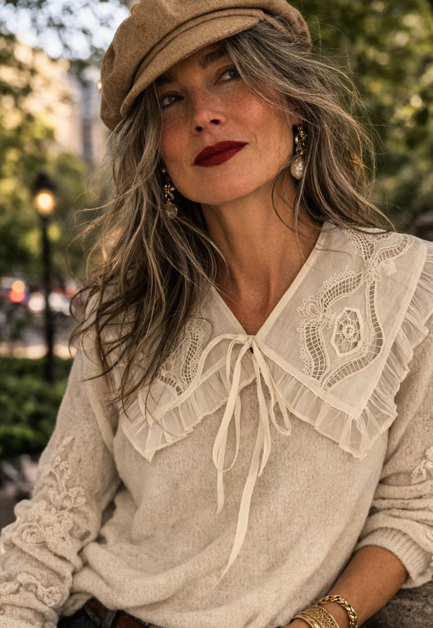 A woman wearing a cream-colored knit sweater with a large ruffled lace collar by the Curated Collar awith bow tie detail, paired with a tan beret, brown belt, and blue jeans, posing outdoors.