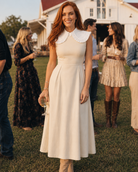 Woman in a cream colored dress with white ruffle collar dickey standing outdoors with people in the background 
