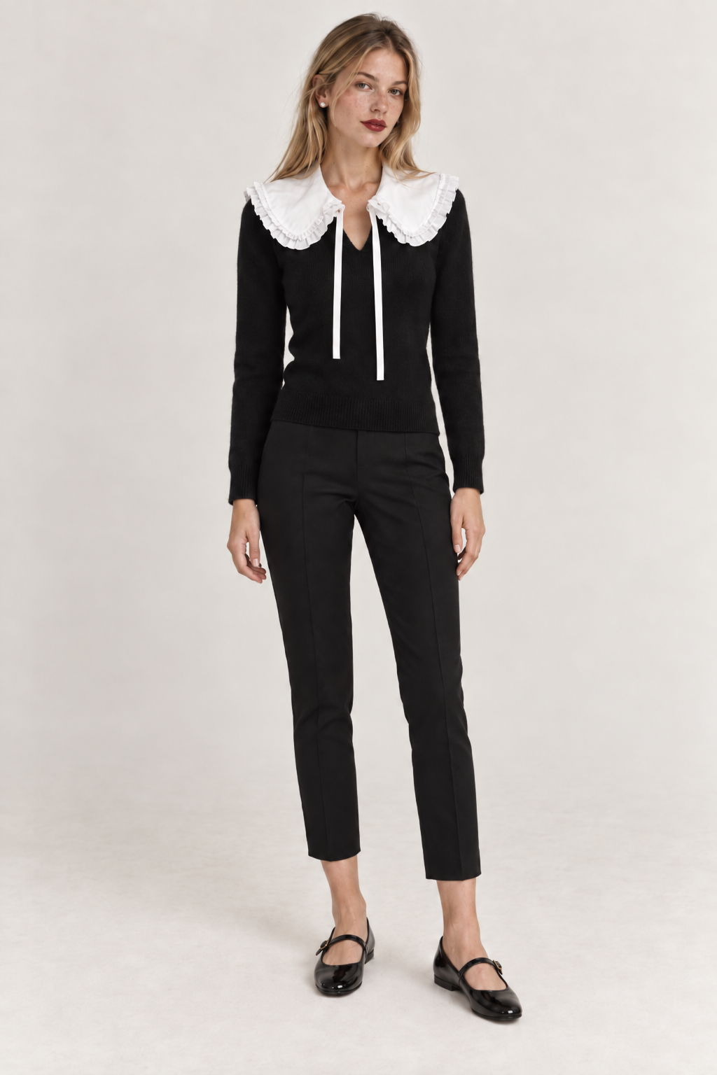 A woman modeling a black knit long-sleeve top with a large white lace Peter Pan collar from the Curated Collar and matching white drawstrings, paired with black cropped trousers and black Mary Jane shoes, against a light gray studio background.