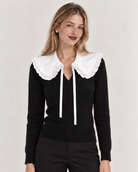 A woman wearing a black ribbed knit sweater with a large white ruffled Peter Pan collar from Curated Collar of Nashville and white drawstrings, paired with black pants, against a light gray studio background.